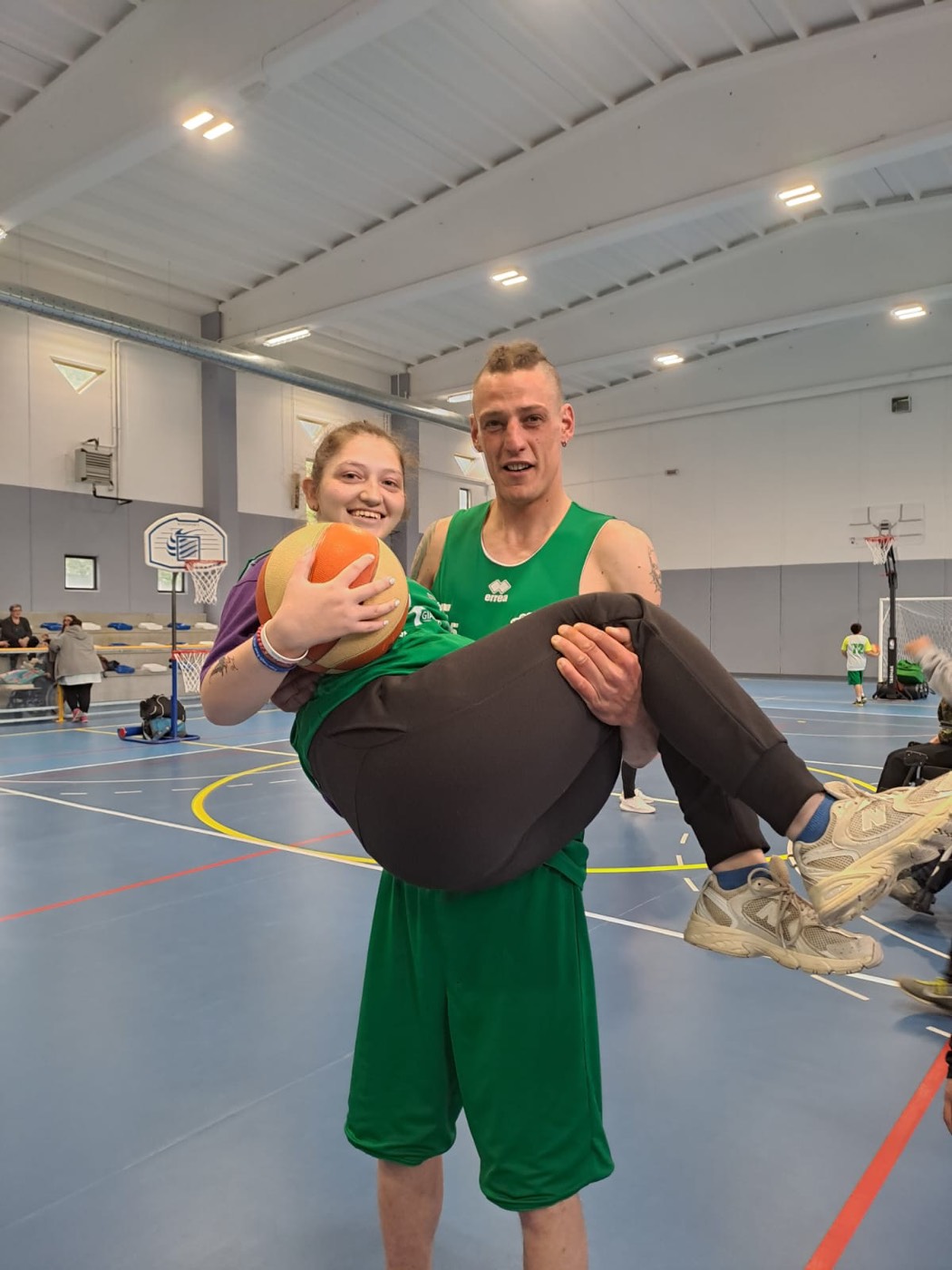 Un giocatore in maglia verde tiene in braccio, scherzando, una compagna di squadra che sorride stringendo il pallone a bordo campo.