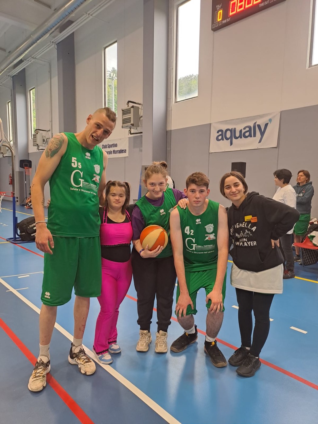Cinque fra giocatori e giocatrici in maglia verde posano sorridenti con il pallone sotto il tabellone del punteggio.